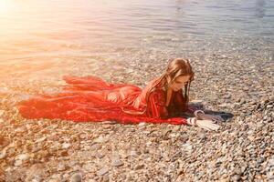 femme Voyage mer. content touristique dans rouge robe prendre plaisir prise image en plein air pour souvenirs. femme voyageur posant dans mer plage, entouré par volcanique montagnes, partage Voyage aventure périple photo