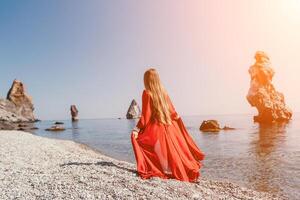 femme Voyage mer. content touristique dans rouge robe prendre plaisir prise image en plein air pour souvenirs. femme voyageur posant sur le Roche à mer baie entouré par volcanique montagnes, partage Voyage aventure périple photo