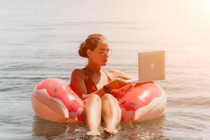 femme pigiste travaux sur portable nager dans mer sur rose gonflable anneau. content touristique flottant sur gonflable Donut et travail sur portable ordinateur dans calme océan. free-lance, éloigné travail nulle part photo