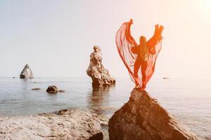 femme Voyage mer. Jeune content femme dans une longue rouge robe posant sur une plage près le mer sur Contexte de volcanique rochers, comme dans Islande, partage Voyage aventure périple photo