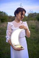 une magnifique femme dans une blanc ancien robe avec une train détient une miroir dans le forme de le lune photo