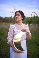 une magnifique femme dans une blanc ancien robe avec une train détient une miroir dans le forme de le lune photo