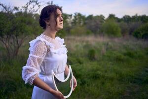 une magnifique femme dans une blanc ancien robe avec une train détient une miroir dans le forme de le lune photo