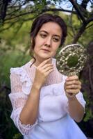 magnifique femme dans blanc ancien robe avec train et en portant ancien miroir dans printemps jardin à le coucher du soleil photo