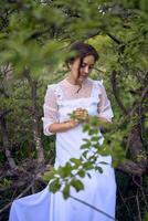 une magnifique femme dans une blanc ancien robe avec une train est prier tandis que séance sur une déchue arbre photo