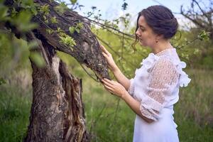 une magnifique femme dans une blanc ancien robe avec une train est caressant une arbre endommagé par une orage photo