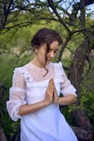 une magnifique femme dans une blanc ancien robe avec une train est prier tandis que séance sur une déchue arbre photo