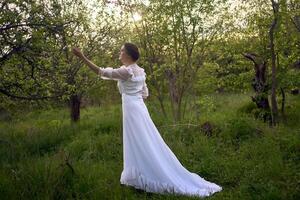 magnifique femme dans blanc ancien robe avec train dans printemps jardin à le coucher du soleil photo