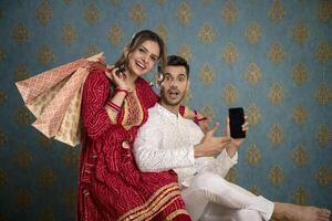 dans de face de le caméra, un attrayant couple habillé dans traditionnel Indien tenue détient une mobile téléphone et achats Sacs photo