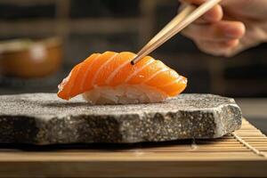 une main en portant baguettes cueillette en haut une pièce de Sushi de une pierre plaque. photo