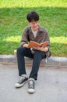 une positif asiatique Masculin Université étudiant est assis sur le rue dans le Campus parc en train de lire une livre. photo