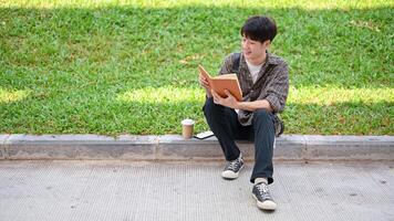 une positif asiatique Masculin Université étudiant est assis sur le rue dans le Campus parc en train de lire une livre. photo