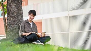 une content, détendu asiatique Masculin Université étudiant jouit en train de lire une livre en dessous de le arbre dans le Campus parc. photo