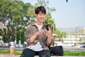 une de bonne humeur Jeune asiatique Masculin Université étudiant est assis sur une pierre banc dans une parc, en utilisant le sien téléphone intelligent. photo