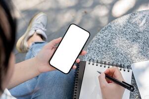 une femme en utilisant sa téléphone intelligent et prise Remarques dans sa carnet tandis que séance à un Extérieur tableau. photo