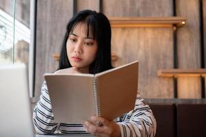 une sérieux asiatique femme est se concentrer sur en train de lire une livre et travail à distance à une café magasin. photo