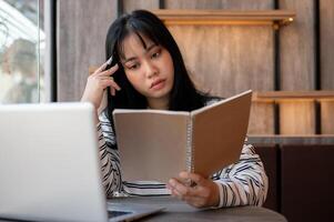 une sérieux asiatique femme est se concentrer sur en train de lire une livre et travail à distance à une café magasin. photo