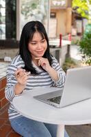 une content asiatique femme dans décontractée vêtements est travail à distance à une café dans le ville. photo