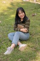 une positif Jeune asiatique femme séance sur le herbe dans une parc avec sa agenda. photo