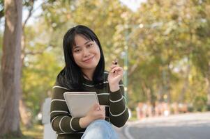 une jolie asiatique femme est séance sur une banc dans une vert parc avec sa carnet de notes, en gardant sa agenda. photo