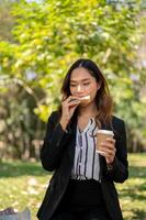 une femme d'affaires est ayant une rapide petit déjeuner dans une ville parc, en mangeant une sandwich et en buvant café. photo