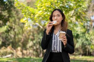 une femme d'affaires est ayant une rapide petit déjeuner dans une ville parc, en mangeant une sandwich et en buvant café. photo