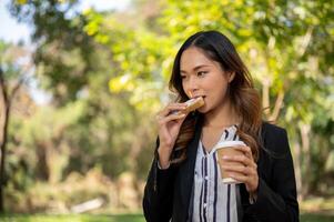 une femme d'affaires est ayant une rapide petit déjeuner dans une ville parc, en mangeant une sandwich et en buvant café. photo