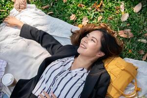 une magnifique asiatique femme d'affaires est mensonge sur une pique-nique tapis, relaxant dans une Publique parc après travail. photo