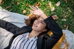 une magnifique asiatique femme d'affaires est mensonge sur une pique-nique tapis, relaxant dans une Publique parc après travail. photo