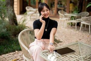 un attrayant asiatique femme est assis à un Extérieur table dans une jardin de une café parlant sur le téléphone. photo