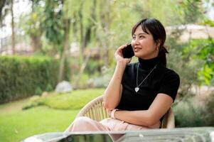 un attrayant asiatique femme est assis à un Extérieur table dans une jardin parlant sur le téléphone avec quelqu'un. photo