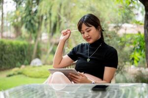 une beau, réfléchi asiatique femme est assis à un Extérieur table dans une jardin en utilisant sa numérique tablette. photo