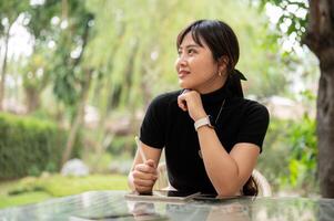 un attrayant asiatique femme est assis à un Extérieur table dans une jardin ou arrière-cour en utilisant sa numérique tablette. photo