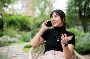 une positif asiatique femme relaxant dans une vert jardin, séance à une table et parlant sur le téléphone. photo