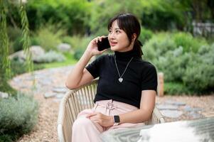 une positif asiatique femme relaxant dans une vert jardin, séance à une table et parlant sur le téléphone. photo