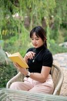 une positif asiatique femme relaxant dans une arrière-cour et en train de lire une livre ou l'écriture sa journal intime à une tableau. photo