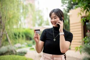 un asiatique femme est appel sa crédit carte ou débit carte appel centre tandis que en marchant dans une jardin. photo