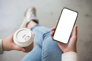 une fermer image de une femme en portant une à emporter café tasse et une téléphone intelligent, séance à l'intérieur. photo