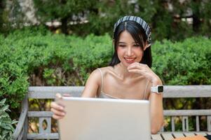 un attractif, positif asiatique femme travail sur sa portable ordinateur à une table dans une vert jardin. photo