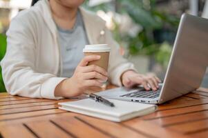 un asiatique femme travail à distance à une café, sirotant café tandis que travail sur sa ordinateur. photo