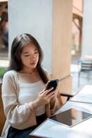 un attrayant asiatique femme est se concentrer sur en utilisant sa téléphone intelligent tandis que séance à un Extérieur tableau. photo