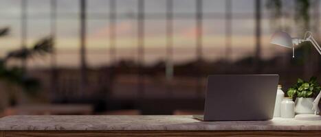 une portable ordinateur, une table lampe, et une mis en pot plante sont sur une table dans une moderne foncé Bureau à le coucher du soleil photo