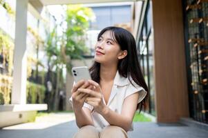 un attrayant asiatique femme est rêverie à propos sa content la vie tandis que en utilisant sa téléphone intelligent dehors. photo