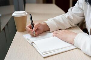 une tondu image de une femme est assis dans une café boutique, l'écriture quelque chose dans une livre ou en gardant sa agenda. photo