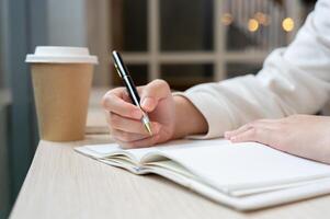 une tondu image de une femme est assis dans une café boutique, l'écriture quelque chose dans une livre ou en gardant sa agenda. photo