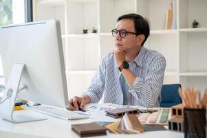 un expert asiatique Sénior Masculin intérieur designer est concentré sur vérification le conception sur le sien ordinateur. photo