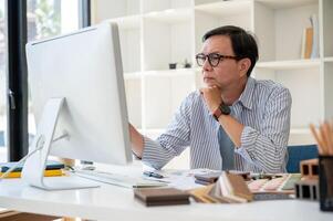 un expert asiatique Sénior Masculin intérieur designer est concentré sur vérification le conception sur le sien ordinateur. photo