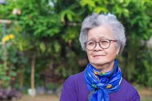 portrait de un personnes âgées femme avec court gris cheveux, souriant, bras franchi, et à la recherche à le caméra tandis que permanent dans une jardin. concept de vieilli gens et soins de santé photo