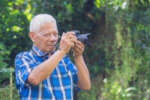 Sénior homme tournage photo par une numérique caméra dans le jardin. un personnes âgées homme porte une bleu chemise, content lorsque en utilisant une caméra. concept de vieilli gens et la photographie