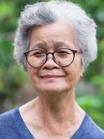 un personnes âgées asiatique femme avec court blanc cheveux souriant et à la recherche à le caméra tandis que permanent dans une jardin. concept de vieilli gens et soins de santé photo